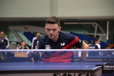 Tom playing table tennis behind net wearing red and blue t-shirt